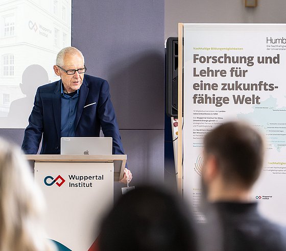 Zu sehen ist Manfred Fischedick vom Wuppertal Institut, der vor einer Gruppe Menschen eine Rede hält. Er steht an einem Podium. Er trägt ein dunkelblaues Sakko und ein blaues Hemd.
