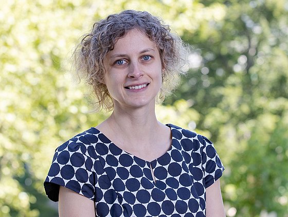 Porträt von Julia Bornhorst vor einem unscharfen Hintergrund mit Bäumen und Büschen. Sie trägt ein Oberteil mit dunkelblauen Kreisen und blickt lächelnd in die Kamera.