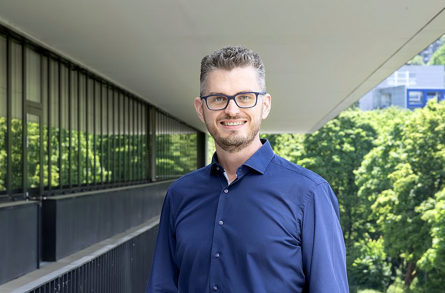 Mann mit Brille, blaues Hemd, kurze Haare, leichter Vollbart, mitteldunkles Haar, lächelt