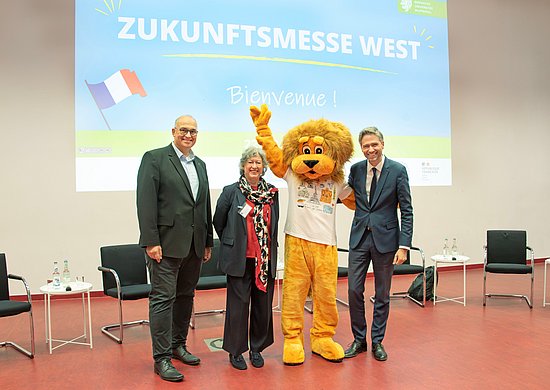 Prof. Dr.-Ing. Peter Gust, Prof. Dr. Andrea von Hülsen-Esch, Xavier Fournier und das Maskottchen der Bergischen Uni - ein lebensgroßer Löwe - stehen vorne im Hörsaal. Im Hintergrund ist die Startseite einer Präsentation über die Zukunftsmesse West an die Wand projiziert.