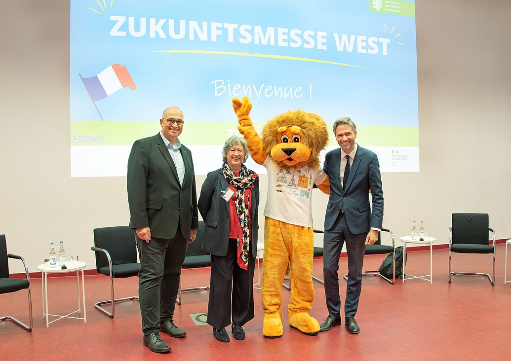 Prof. Dr.-Ing. Peter Gust, Prof. Dr. Andrea von Hülsen-Esch, Xavier Fournier und das Maskottchen der Bergischen Uni - ein lebensgroßer Löwe - stehen vorne im Hörsaal. Im Hintergrund ist die Startseite einer Präsentation über die Zukunftsmesse West an die Wand projiziert.