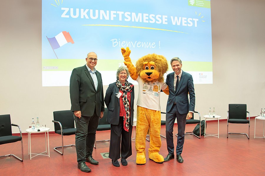 Prof. Dr.-Ing. Peter Gust, Prof. Dr. Andrea von Hülsen-Esch, Xavier Fournier und das Maskottchen der Bergischen Uni - ein lebensgroßer Löwe - stehen vorne im Hörsaal. Im Hintergrund ist die Startseite einer Präsentation über die Zukunftsmesse West an die Wand projiziert.