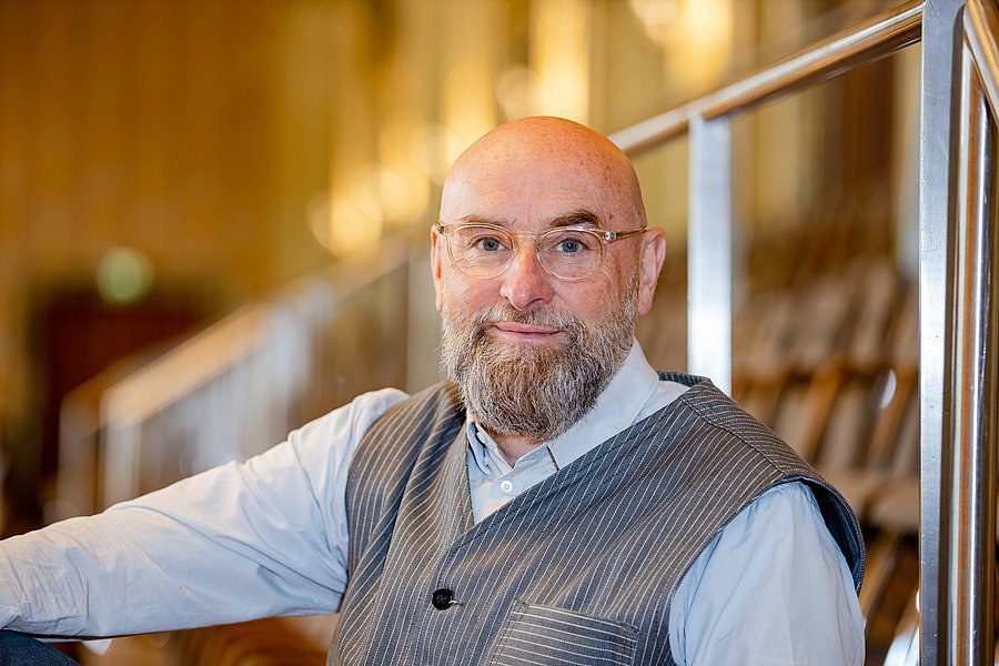 Prof. Topel sitzt auf der Empore in der Historischen Stadthalle in Wuppertal. Er trägt ein helles Hemd mit einer grauen Weste und lächelt in die Kamera. Er trägt eine Brille und einen Vollbart.  