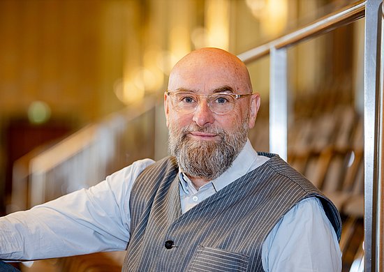 Prof. Topel sitzt auf der Empore in der Historischen Stadthalle in Wuppertal. Er trägt ein helles Hemd mit einer grauen Weste und lächelt in die Kamera. Er trägt eine Brille und einen Vollbart.