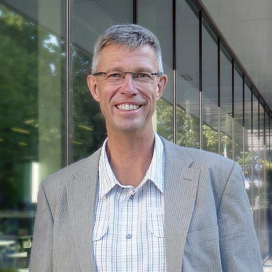 Porträtaufnahme von Prof. Dr. Jürgen Gerlach. Mann mittleren Alters, graues, kurzes Haar, Brille, blickt lächelnd in die Kamera, trägt ein weißes, kariertes Hemd und ein graues Sakko.