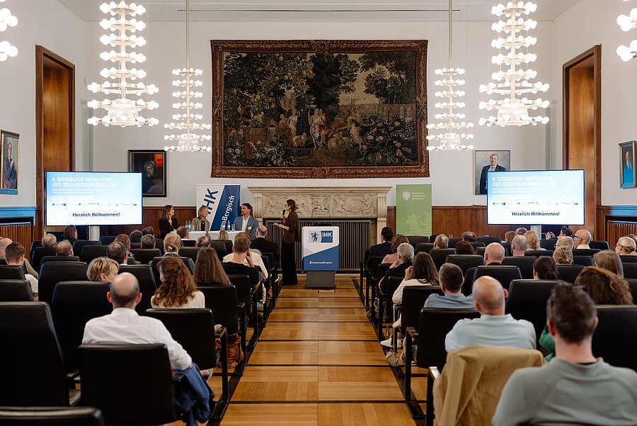 The photograph shows the plenary hall of the Bergisch Chamber of Industry and Commerce. Seated at the front are Miriam Scherff, Mayor of Wuppertal; Prof. Dr Birgitta Wolff, rector of the university of Wuppertal; and Henner Pasch, President of the Chamber of Industry and Commerce, who are talking to one another. The audience can be seen from behind.   