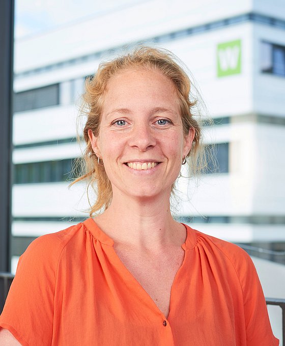 Porträtaufnahme von Marie Cravageot. Eine blonde Frau, die lächelt und ein orangefarbenes Oberteil trägt. Im Hintergrund ist unscharf ein Gebäude der Bergischen Universität zu sehen.