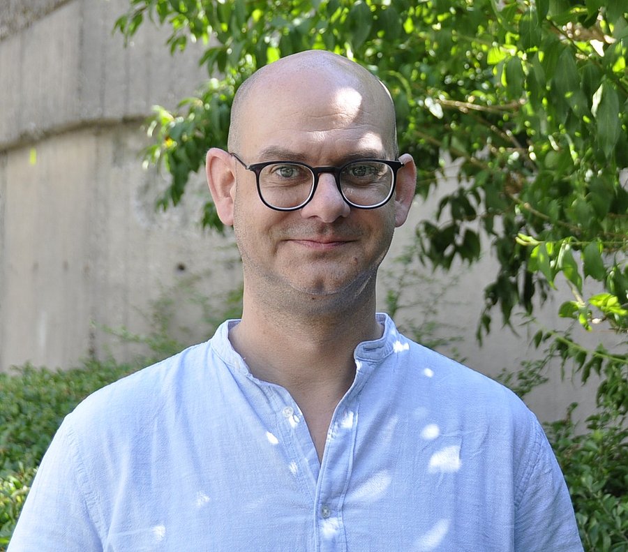 Porträtaufnahme von Christoph Gardian. Mann mit Glatze, trägt eine schwarze Brille und ein hellblaues Hemd. Im Hintergrund sind verschwommen eine Betonwand sowie Blätter eines Baumes zu sehen.