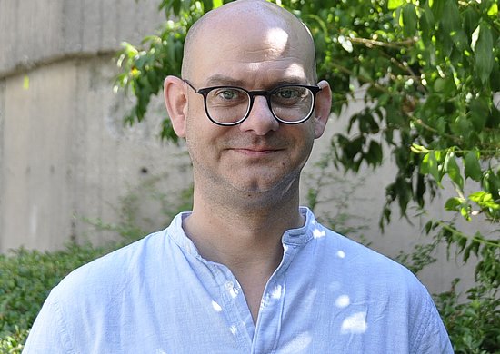 Porträtaufnahme von Christoph Gardian. Mann mit Glatze, trägt eine schwarze Brille und ein hellblaues Hemd. Im Hintergrund sind verschwommen eine Betonwand sowie Blätter eines Baumes zu sehen.