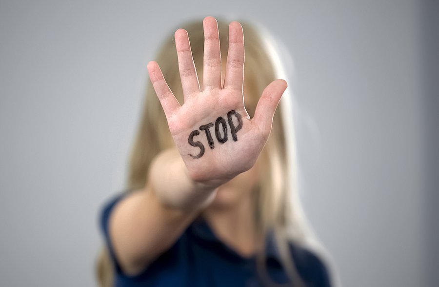 Das Bild zeigt eine Person mit langen, blonden Haaren, die ihre Hand mit gespreizten Fingern in den Vordergrund hält. Auf der Handfläche steht das Wort „STOP“ in schwarzer Schrift. Das Gesicht der Person ist unscharf und verdeckt, während der Fokus auf der Hand liegt. Der Hintergrund ist neutral und verschwommen. Das Bild vermittelt eine starke Botschaft gegen Gewalt oder unerwünschtes Verhalten.