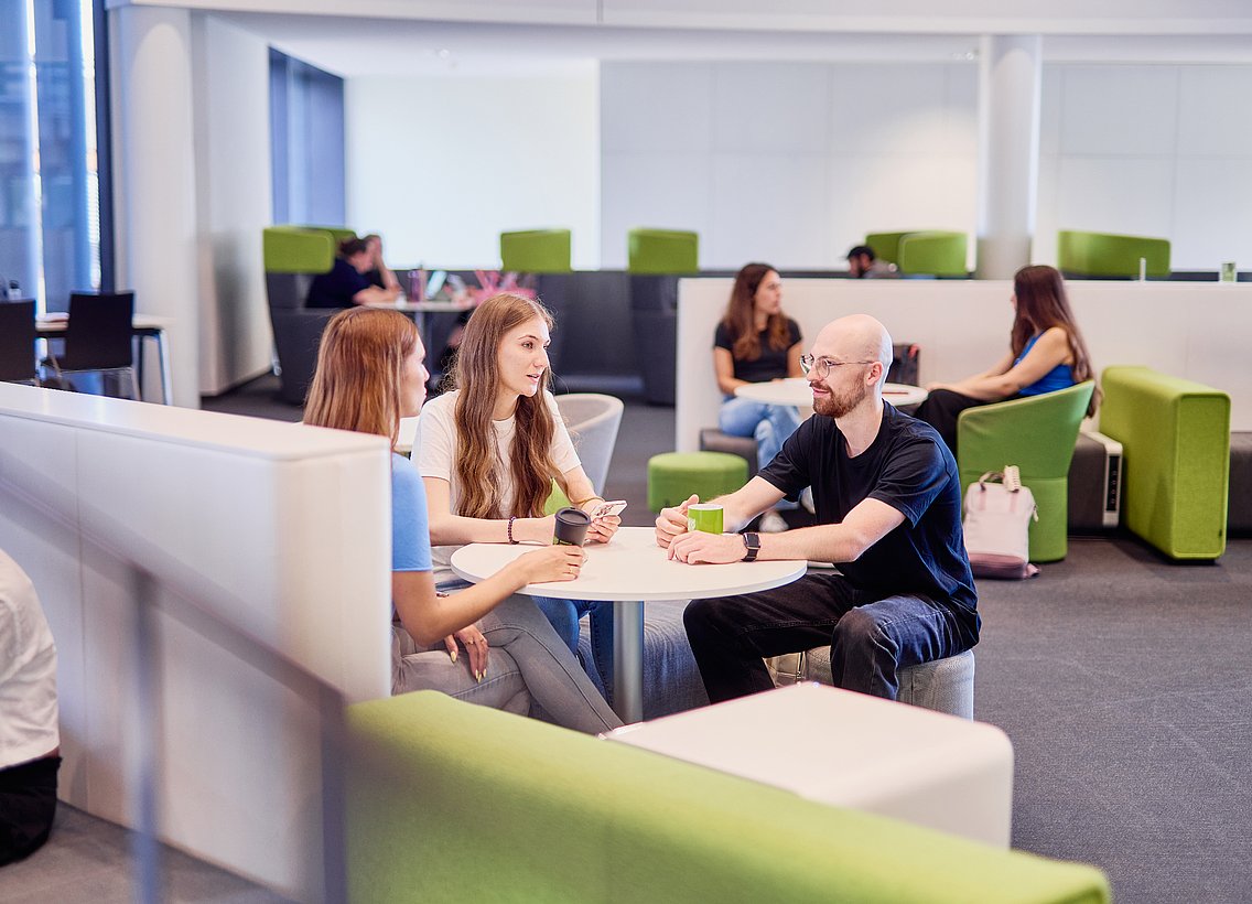 Das Bild zeigt zwei Frauen und einen Mann, die an einem Tisch in der Universitätsbibliothek sitzen und sich miteinander unterhalten. 