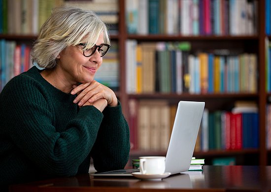 Eine ältere Frau mit kurzen grauen Haaren und Brille sitzt an einem Tisch vor einem Bücherregal. Sie blickt lächelnd auf einen geöffneten Laptop. Neben dem Laptop stehen mehrere Bücher und eine Tasse Kaffee.