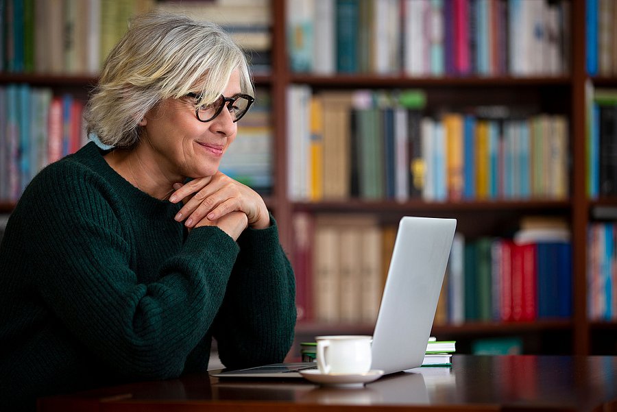Eine ältere Frau mit kurzen grauen Haaren und Brille sitzt an einem Tisch vor einem Bücherregal. Sie blickt lächelnd auf einen geöffneten Laptop. Neben dem Laptop stehen mehrere Bücher und eine Tasse Kaffee.