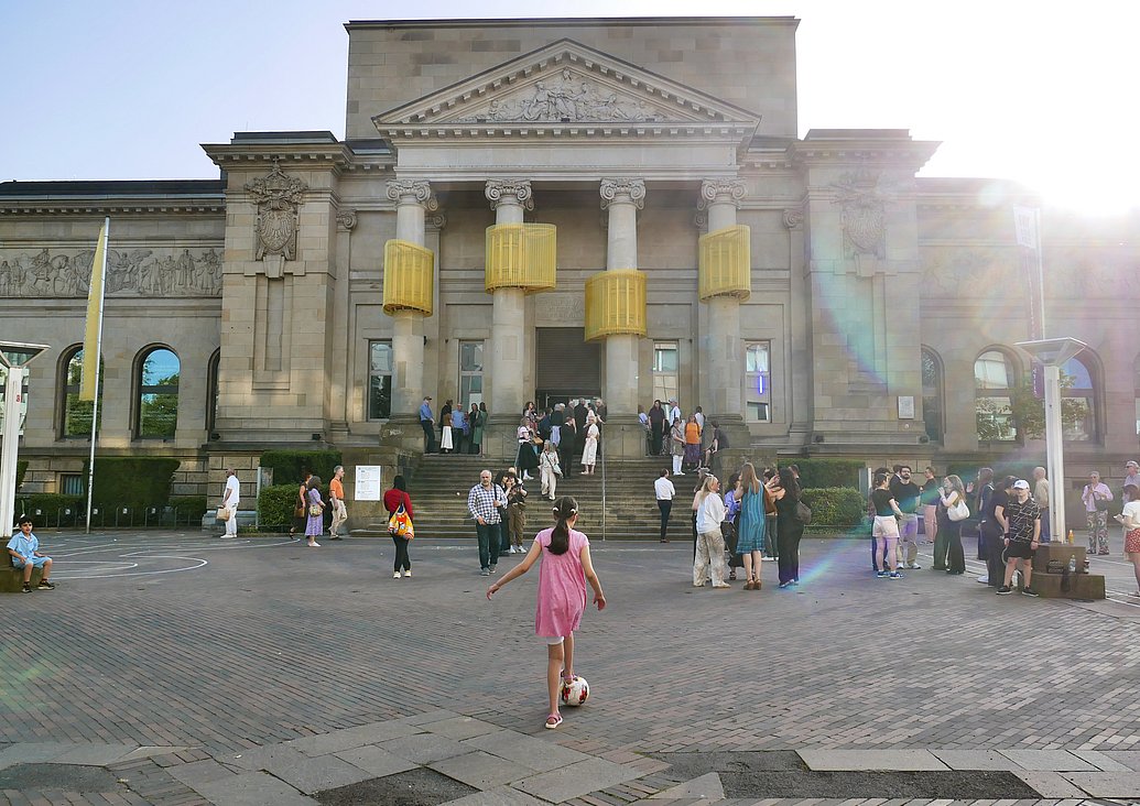 Frontalaufnahme der Kunsthalle Barmen inklusive Vorplatz. Auf dem Vorplatz sind zahlreiche Menschen zu sehen, ein Mädchen in der Mitte des Bildes spielt Fußball. Andere Menschen stehen in Gruppen zusammen und unterhalten sich