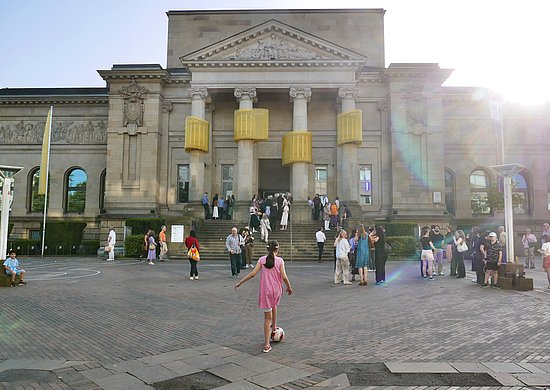Frontalaufnahme der Kunsthalle Barmen inklusive Vorplatz. Auf dem Vorplatz sind zahlreiche Menschen zu sehen, ein Mädchen in der Mitte des Bildes spielt Fußball. Andere Menschen stehen in Gruppen zusammen und unterhalten sich