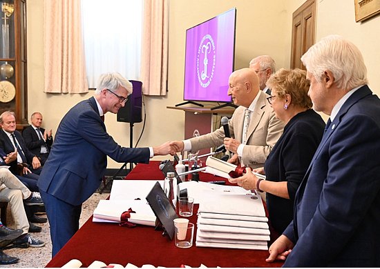 Innenraum der Akademie, links stehen vier Vertreter*innen der Akademie an einem langen  Tisch mit roter Tischplatte, Prof Johrendt rechts vom Tisch nimmt die Urkunde entgegen und schüttelt einer Person die Hand, am linken Bildrand weiteres Publikum sitzend angdeutet.