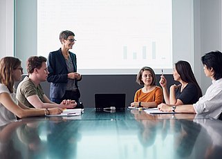 Eine Besprechungssituation, eine Person steht vor einer Leinwand. Fünf Personen sitzen an einem Tisch, eine davon spricht und gestikuliert. 