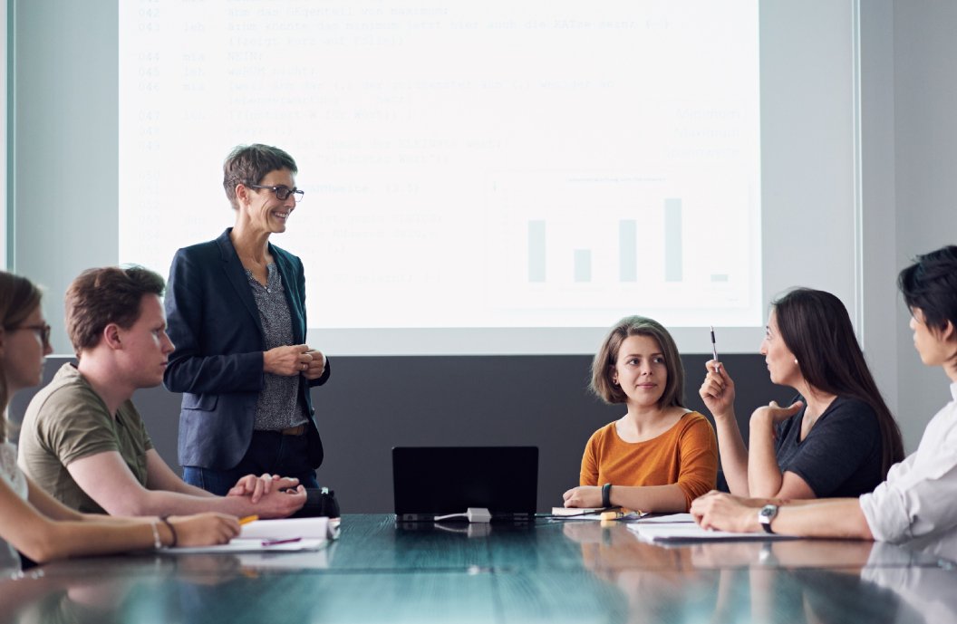 Eine Besprechungssituation, eine Person steht vor einer Leinwand. Fünf Personen sitzen an einem Tisch, eine davon spricht und gestikuliert. 