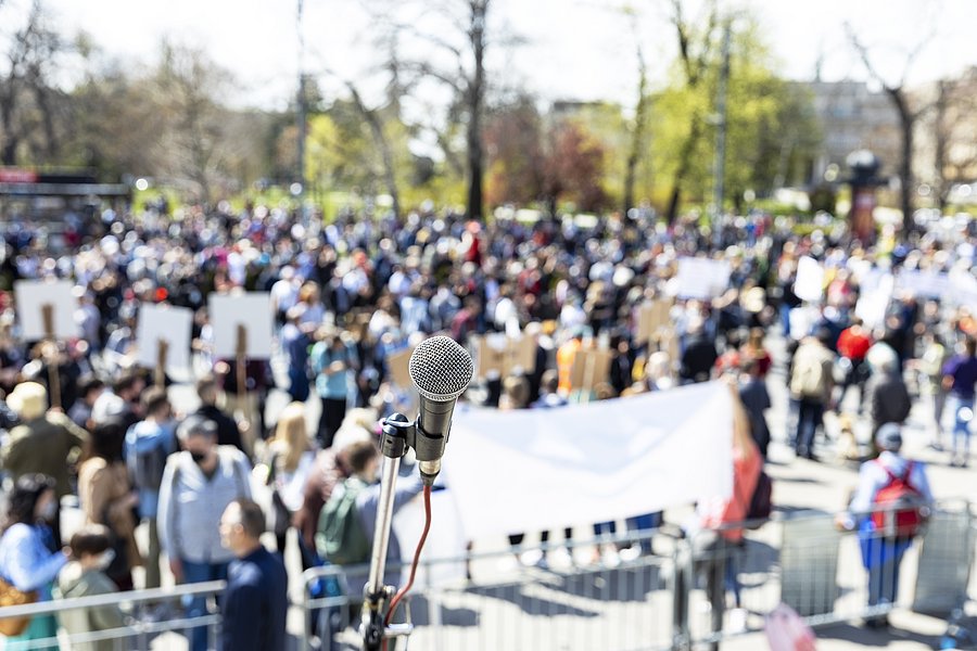 Im Hintergrund Menschenmenge, die sich für eine Kundgebung versammelt, halten teilweise Schilder hoch. Im Vordergrund ein Mikrofon an einem Ständer.
