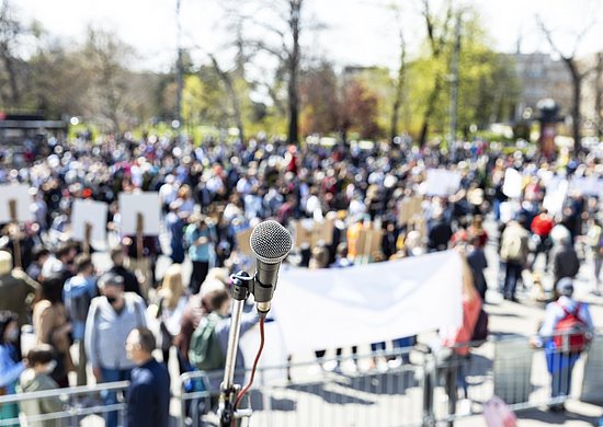 Im Hintergrund Menschenmenge, die sich für eine Kundgebung versammelt, halten teilweise Schilder hoch. Im Vordergrund ein Mikrofon an einem Ständer.