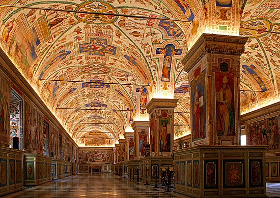 Prunkvoll ausgemalter Bibliothekssaal im Vatikan mit hohen, reich verzierten Gewölben, Wandgemälden und holzvertäfelten Bücherregalen. Eine lange, von Säulen gesäumte Galerie führt in die Tiefe des Saals.