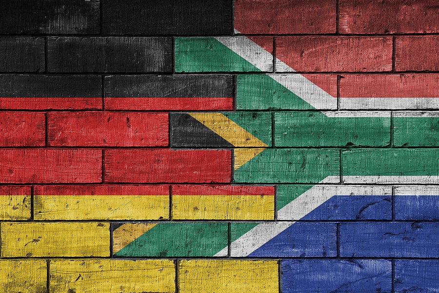 The picture shows a creative representation of the German and South African flags painted on a brick wall. The two flags are fused together so that they merge seamlessly. The rough, textured surface of the bricks gives the picture a rustic and artistic look.