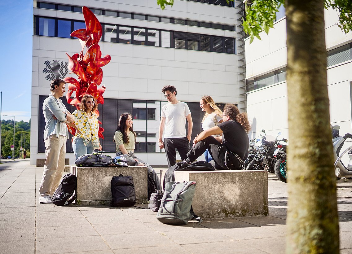 Studierende sitzen vor dem Unigebäude V/W und unterhalten sich, Sonne scheint, blauer Himmel
