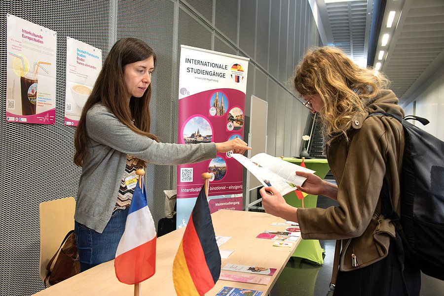 Eine brünette Frau steht hinter einem Tisch und erklärt einer Frau auf der anderen Seite des Tisches etwas, was in einem Flyer steht, den diese in der Hand hält.