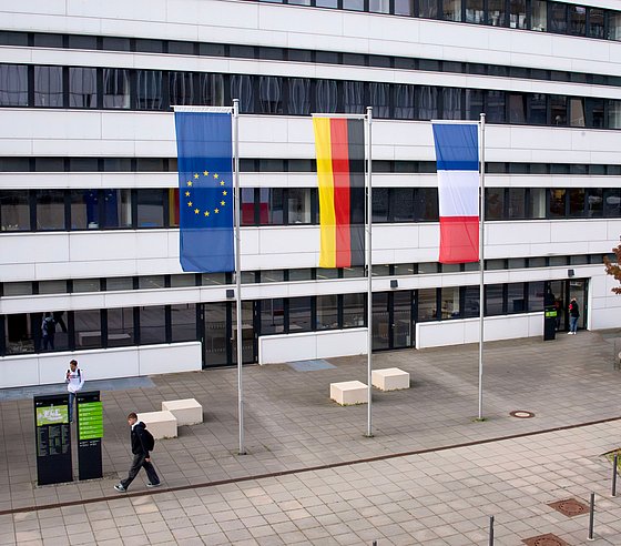 Vor einem Universitätsgebäude stehen drei Fahnenmasten. Daran sind jeweils die Europaflagge, die deutsche sowie die französische Flagge gehisst.