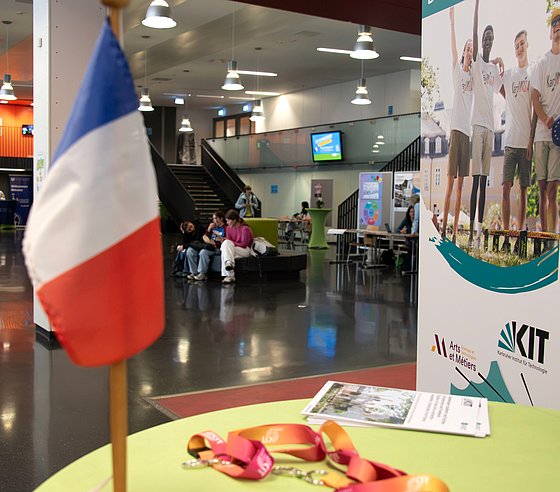 Das Foyer von Gebäude K, fotografiert von einem Stehtisch aus. Rechts steht ein Roll-up einer der ausstellenden Institutionen. Im Vordergrund ist eine französische Tischfahne zu sehen.