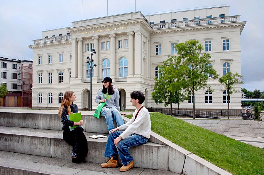 Drei junge Personen sitzen auf den Stufen vor der ehemaligen Bundesbahndirektion in Wuppertal und unterhalten sich. Links sitzt eine junge Frau, sie ist schwart gekleidet und hat lange Haare, in der Hand hält sie einen Ordner. In der Mitte sitzt eine junge Frau mit braunen Haaren. Sie trägt einen Hut, eine blaue Jeans und einen grauen Pullover. Recht sitzt ein junger Mann mit dunklen Haaren. Er trägt braune Schuhe, eine blaue Jeans und ein helles Oberteil.