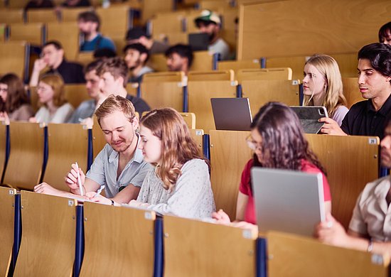 Das Foto zeigt Studierende in einem Hörsaal der Bergischen Universität. Manche haben einen Laptop vor sich stehen, andere nutzen Stift und Papier. Sie folgen der Vorlesung, zwei der Studierenden unterhalren sich miteinander. 