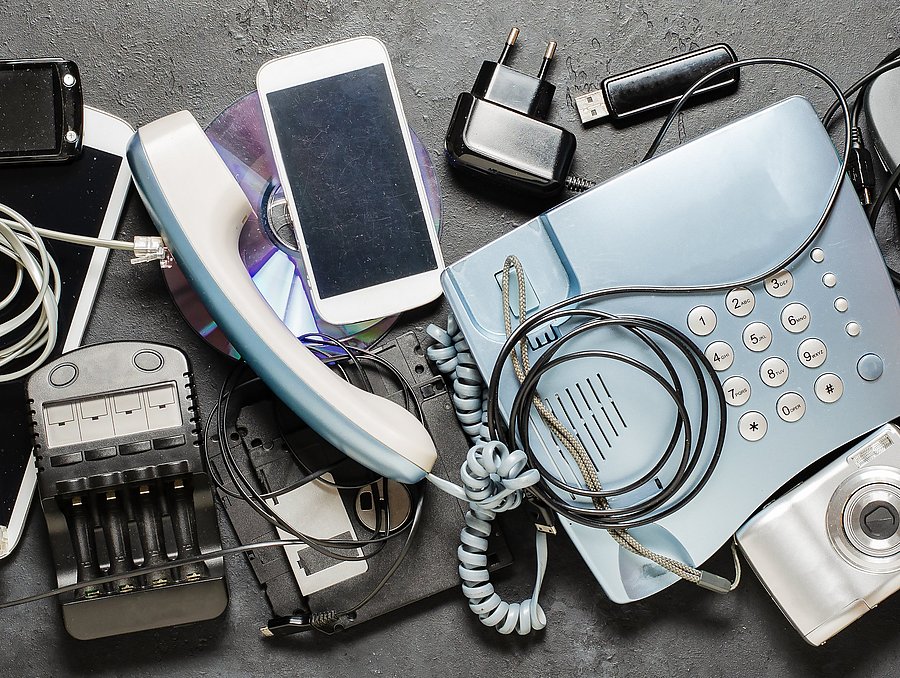 Auf einer dunklen Oberfläche liegen verschiedene ausgediente Elektrogeräte und Zubehörteile, darunter eine Tastatur, mehrere Mobiltelefone, ein Festnetztelefon, eine Computermaus, eine Digitalkamera, Ladegeräte, Kabel und ein Taschenrechner.