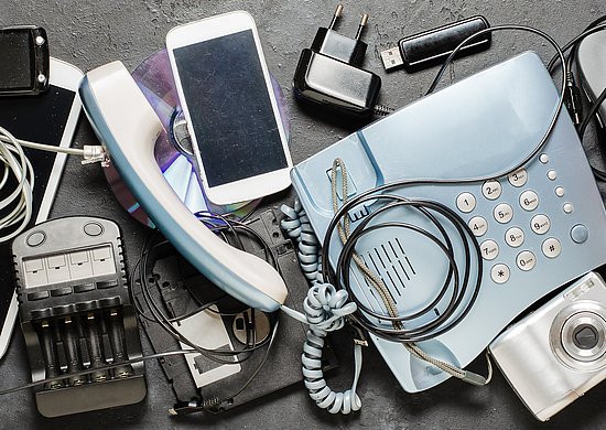 Auf einer dunklen Oberfläche liegen verschiedene ausgediente Elektrogeräte und Zubehörteile, darunter eine Tastatur, mehrere Mobiltelefone, ein Festnetztelefon, eine Computermaus, eine Digitalkamera, Ladegeräte, Kabel und ein Taschenrechner.