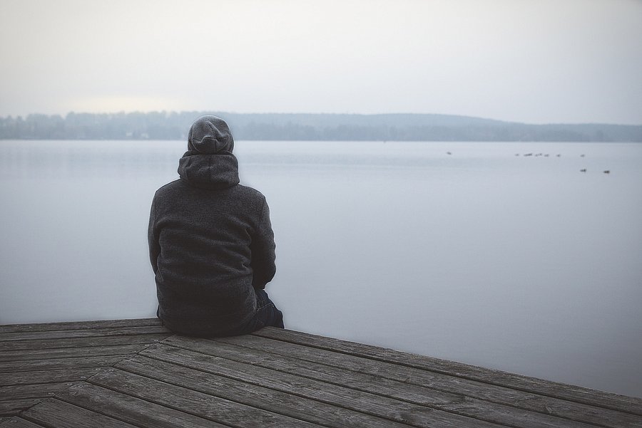 Eine Person sitzt mit dem Rücken zur Kamera allein auf einem Holzsteg und blickt auf einen ruhigen, nebligen See. Am gegenüberliegenden Ufer ist eine bewaldete Landschaft zu sehen.