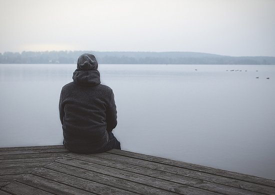 Eine Person sitzt mit dem Rücken zur Kamera allein auf einem Holzsteg und blickt auf einen ruhigen, nebligen See. Am gegenüberliegenden Ufer ist eine bewaldete Landschaft zu sehen.
