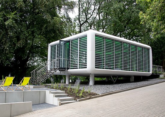 Außenansicht des neu eröffneten „Orts der Stille“ auf dem Campus Grifflenberg der Bergischen Universität Wuppertal: Ein moderner, pavillonartiger Bau mit großen Glasflächen und weißen Lamellen steht erhöht in der Nähe von Bäumen. Eine Treppe führt zum Eingang; davor befinden sich Sitzstufen und zwei gelbe Liegestühle.
