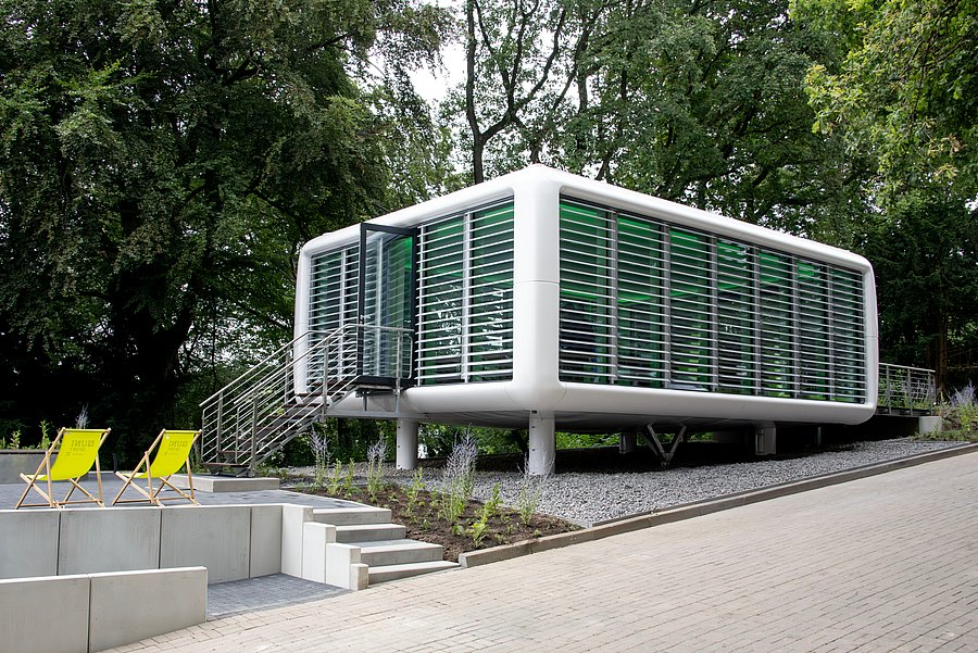 Außenansicht des neu eröffneten „Orts der Stille“ auf dem Campus Grifflenberg der Bergischen Universität Wuppertal: Ein moderner, pavillonartiger Bau mit großen Glasflächen und weißen Lamellen steht erhöht in der Nähe von Bäumen. Eine Treppe führt zum Eingang; davor befinden sich Sitzstufen und zwei gelbe Liegestühle.