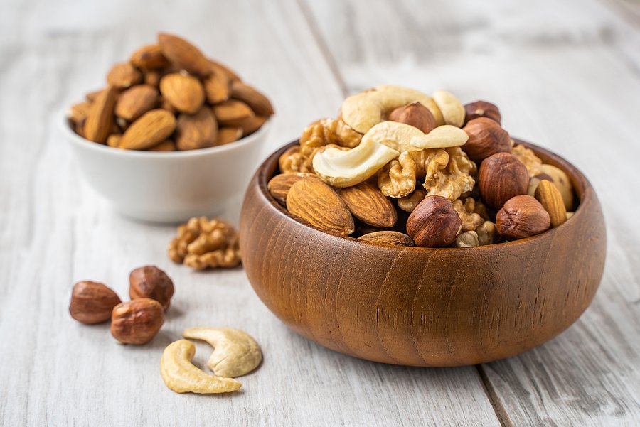Verschiedene Nüsse wie Mandeln, Walnüsse, Haselnüsse und Cashewkerne liegen in einer Holzschale auf einem hellen Holztisch; im Hintergrund steht eine weitere Schale mit Mandeln.