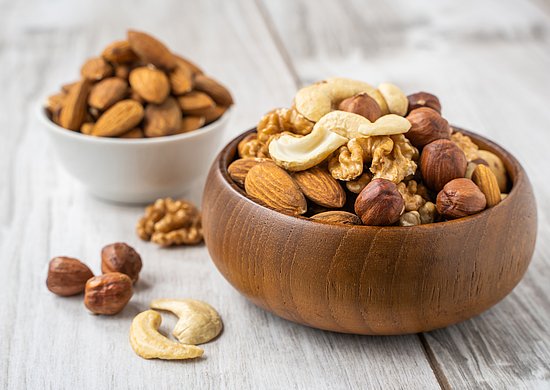 Verschiedene Nüsse wie Mandeln, Walnüsse, Haselnüsse und Cashewkerne liegen in einer Holzschale auf einem hellen Holztisch; im Hintergrund steht eine weitere Schale mit Mandeln.