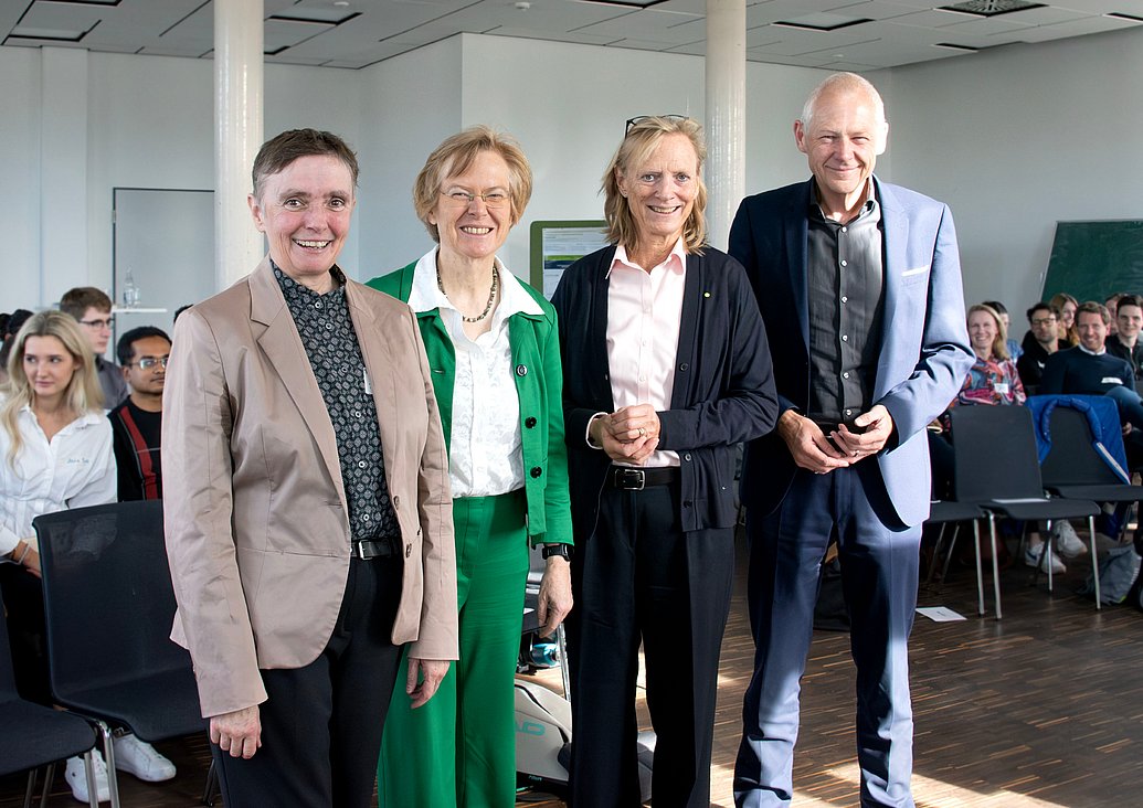 Das Bild zeigt einen Vortragsraum, in dem einige Menschen auf Stühlen sitzen. Im Vordergrund steht eine Gruppe von vier Personen.