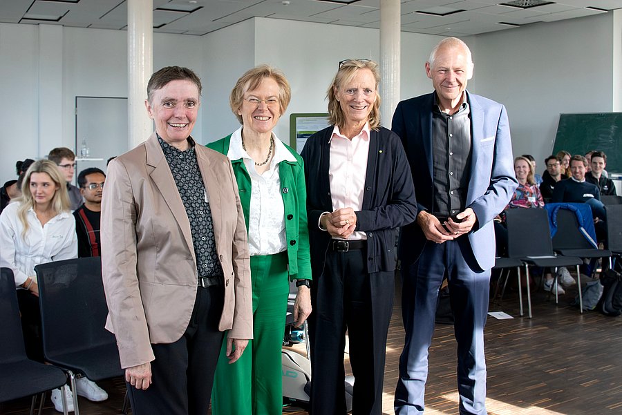 Das Bild zeigt einen Vortragsraum, in dem einige Menschen auf Stühlen sitzen. Im Vordergrund steht eine Gruppe von vier Personen.