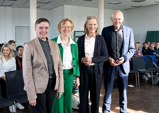 Das Bild zeigt einen Vortragsraum, in dem einige Menschen auf Stühlen sitzen. Im Vordergrund steht eine Gruppe von vier Personen.
