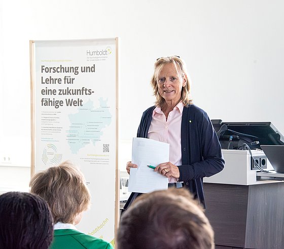 Zu sehen ist Uni-Rektorin Birgitta Wolff, die vor einer Gruppe Menschen spricht. Sie hält ein Blatt Papier in der Hand. Sie trägt ein hellrosa Hemd und eine dunkelblaue Strickjacke.
