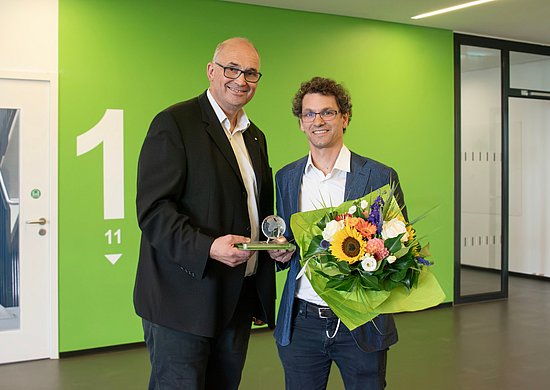 Bei einer Preisverleihung stehen zwei Personen nebeneinander und präsentieren gemeinsam eine gläserne Auszeichnung. Die ausgezeichnete Person hält zusätzlich einen bunten Blumenstrauß.