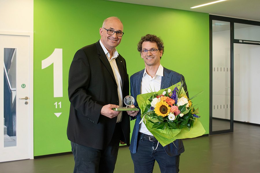 Bei einer Preisverleihung stehen zwei Personen nebeneinander und präsentieren gemeinsam eine gläserne Auszeichnung. Die ausgezeichnete Person hält zusätzlich einen bunten Blumenstrauß.