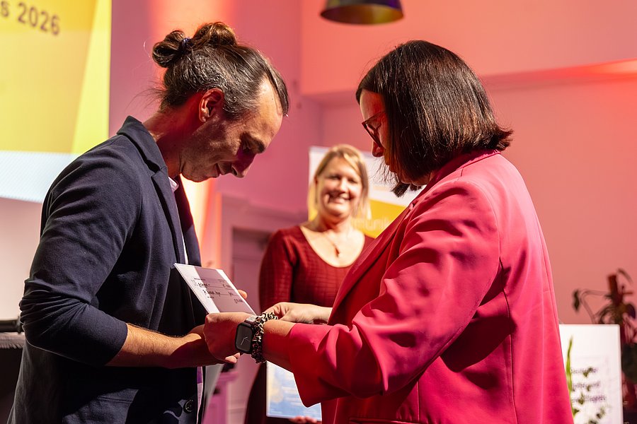 Ein Mann gekleidet in Sakko hält eine Zettel in der Hand, der ihm von einer Frau rechts von ihm überreicht wird. Die Frau trägt einen roten Blazer, beide schauen zur Mitte des Bildes, nicht in die Kamera.