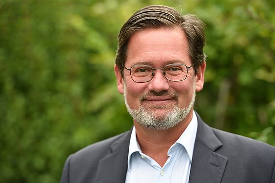 Porträt von Historiker Arne Karsten vor einem unscharfen Hintergrund mit Bäumen und Büschen. Er trägt eine Brille, ein weißes Hemd sowie ein dunkelgraues Sakko und blickt lächelnd in die Kamera.