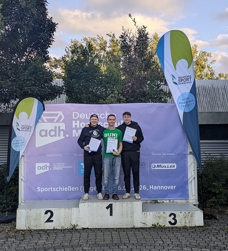 Drei Männer stehen auf dem Siegertreppen eines sportlichen Wettbewerbs und halten ihre Urkunden vor der Brust. Im Untergrund ist ein Banner vom Allgemeinen Deutschen Hochschulsportverband zu sehen. 