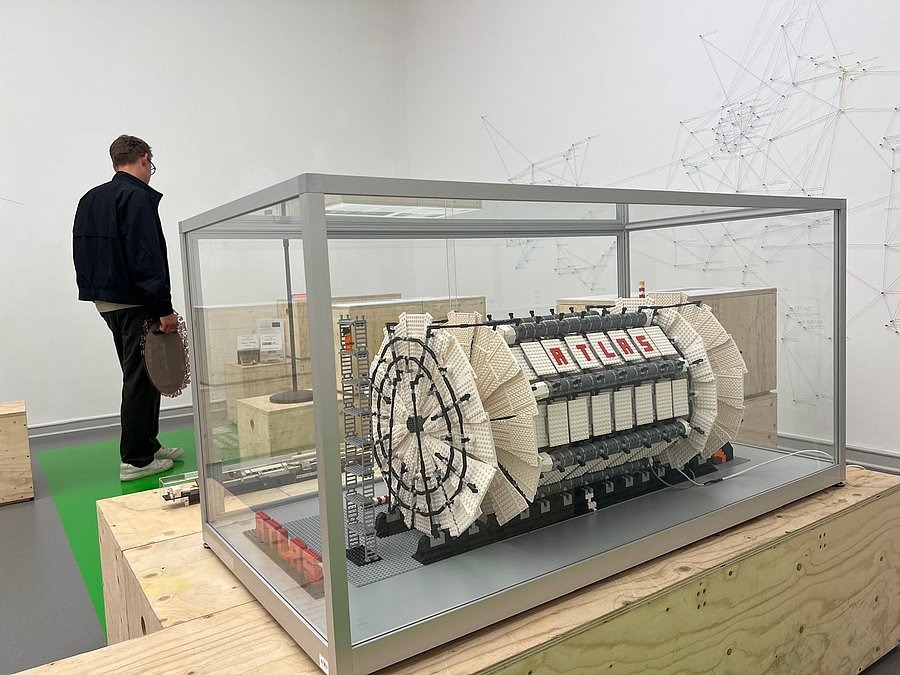 Ein Modell des Teilchendetektors ATLAS aus Lego, das derzeit in der Kunsthalle Barmen ausgestellt wird. Es befindet sich in einem Kasten aus Glas. Im Hintergrund ist ein Mann von hinten zu sehen, der sich ein anderes Ausstellungsobjekt anschaut. 